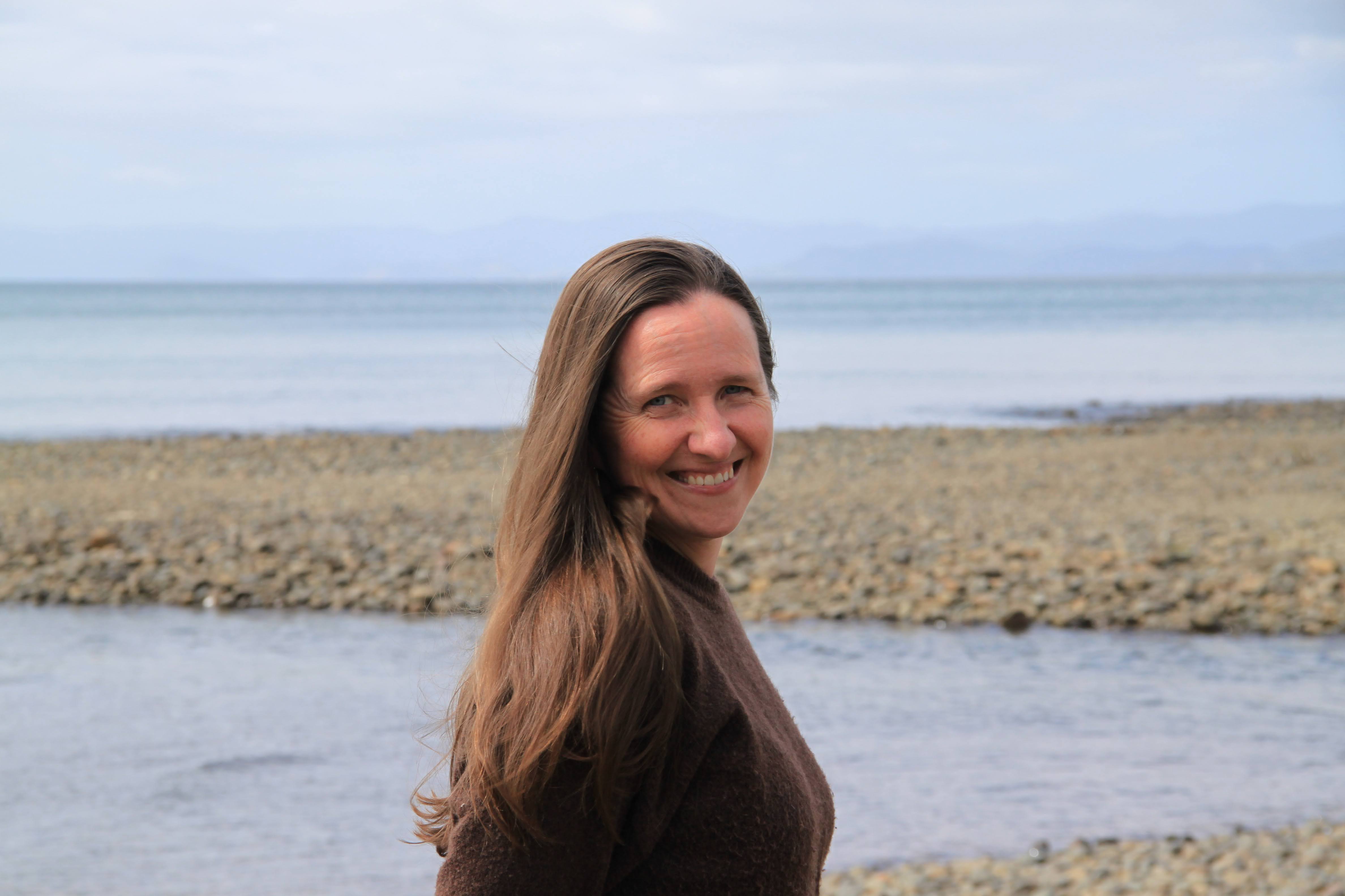 Johanna standing at a pebble beach on a calm overcast day, smiling warmly over her shoulder toward the camera, with sea and distant mountains behind.