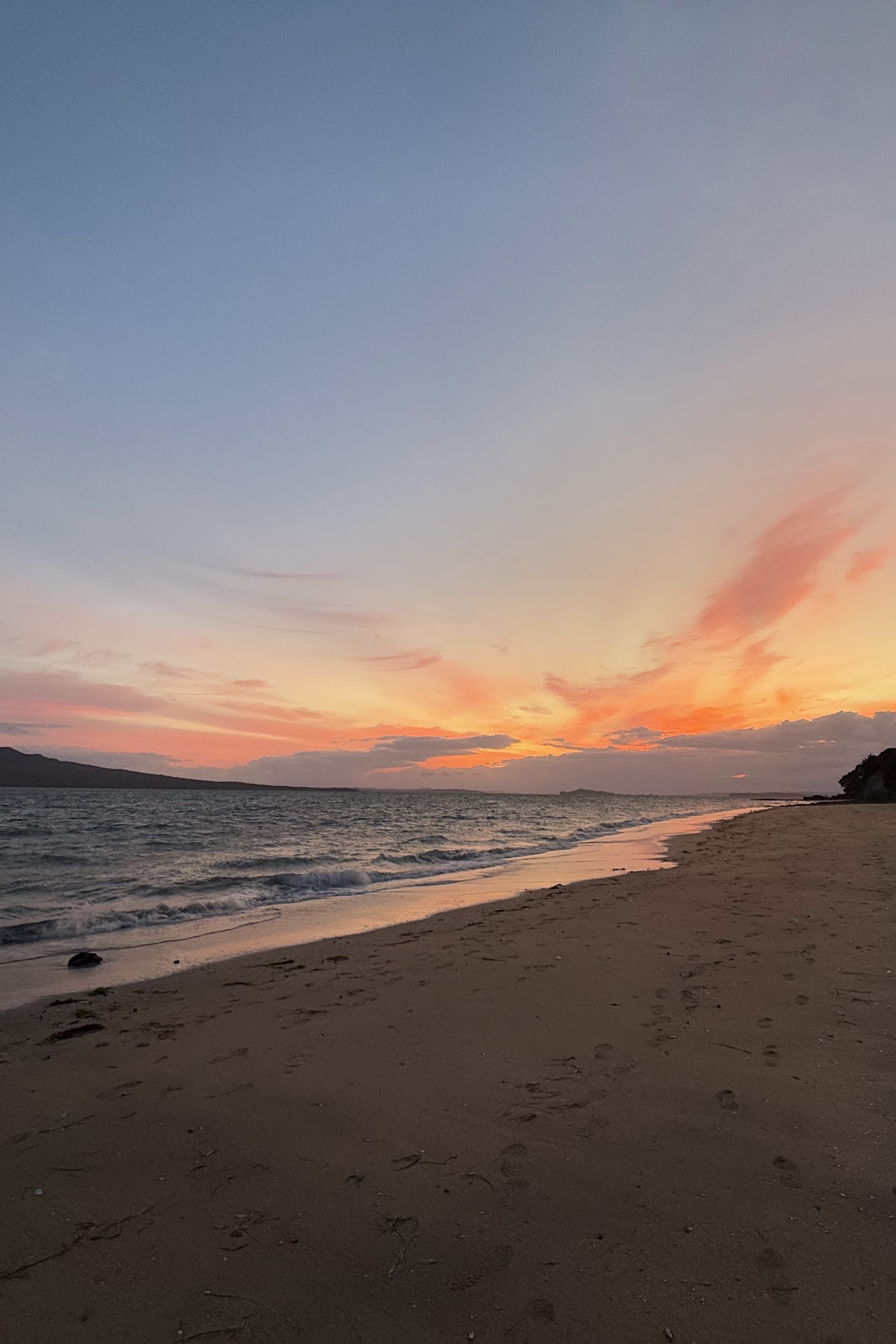 An early dawn beach with orange-tinted sunrise light over calm ocean and dark sand, with a mountain silhouette across the water.