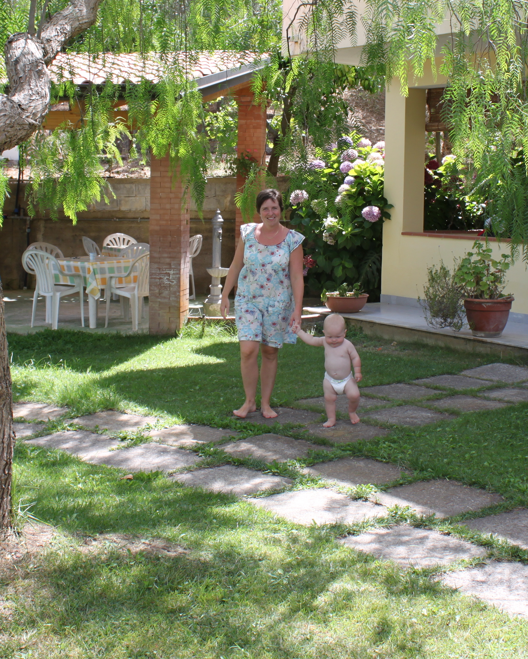 Johanna in a turquoise floral dress stands with her young son Valdemar in a sunny garden courtyard; a weeping tree arches overhead, a stone path winds through the grass, and a small house with potted plants sits behind them.