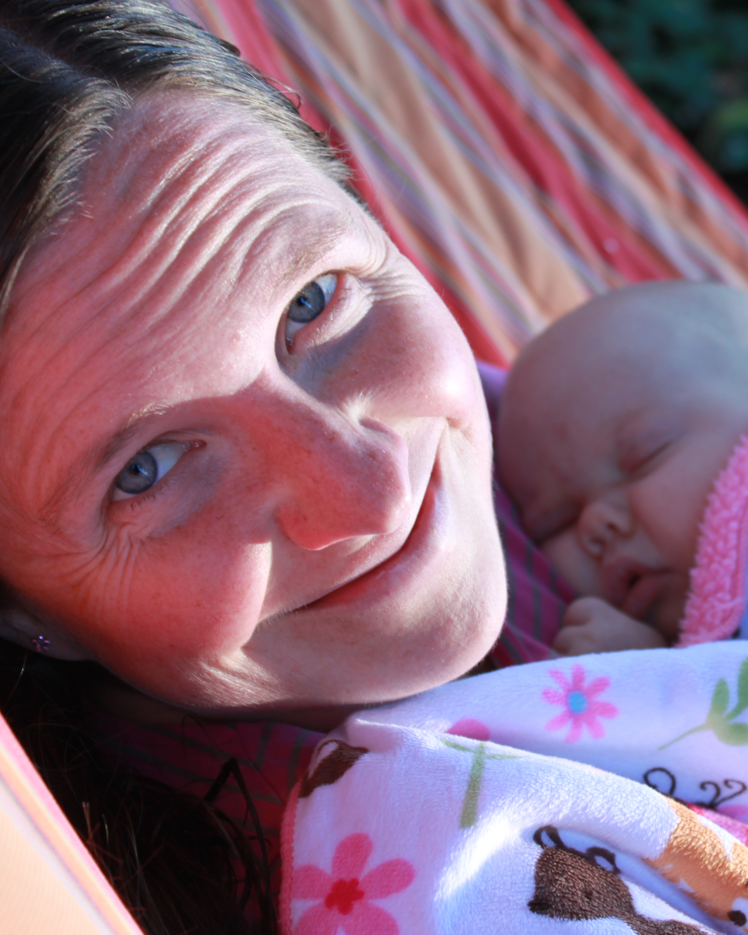 Johanna in a striped hammock holding a sleeping newborn baby, lit by warm late-afternoon sun.