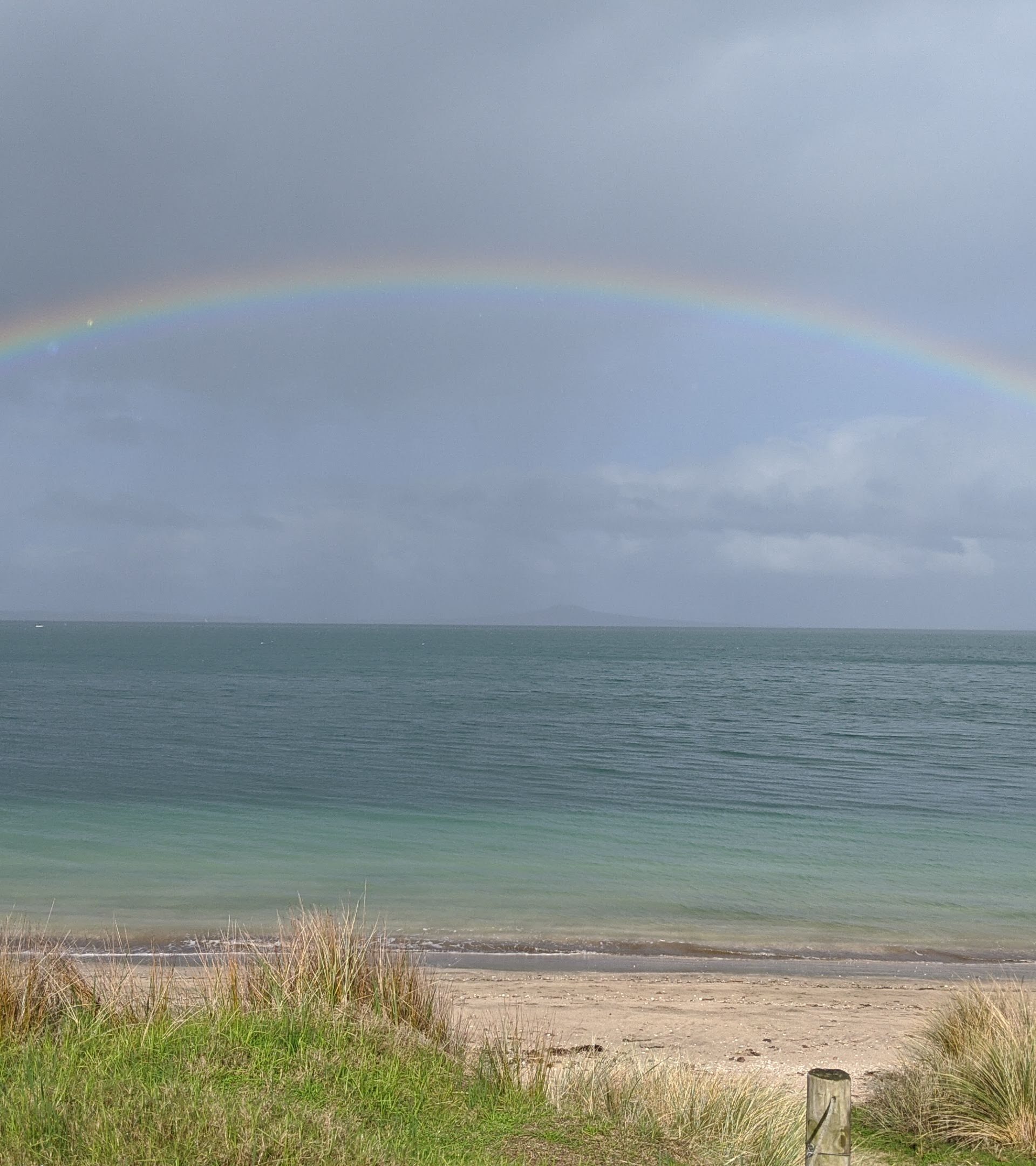 A wide double rainbow arches across a calm ocean and overcast sky, with a small boat near the left shore and a quiet sandy beach in the foreground.