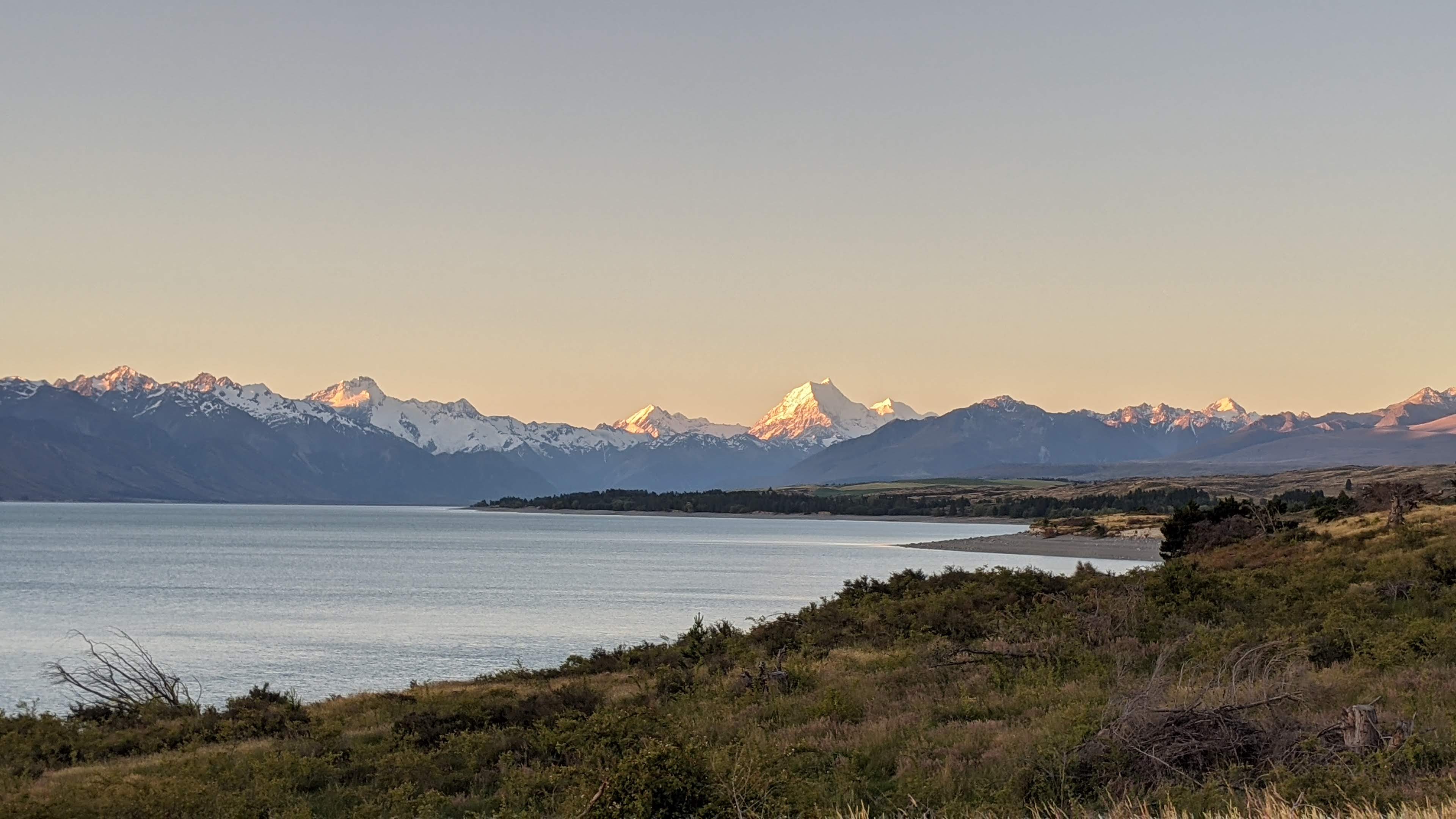 An alpine lake at dawn with golden light on snow-capped mountains across the water; soft warm lighting and muted vegetation along the shoreline.