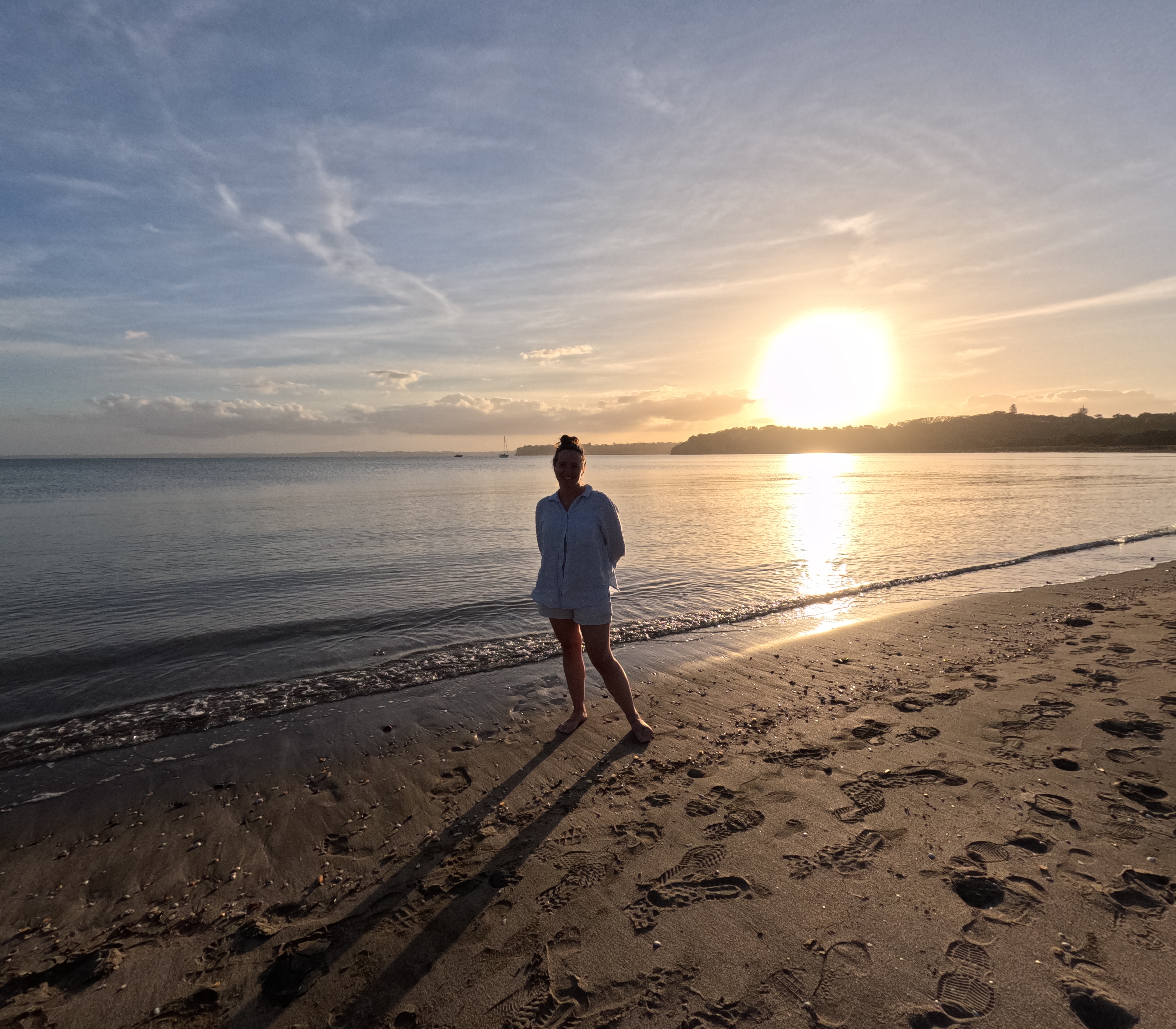 Johanna walks along a sandy beach at sunset, seen from behind as a silhouette against the brilliant setting sun and golden reflections on wet sand.